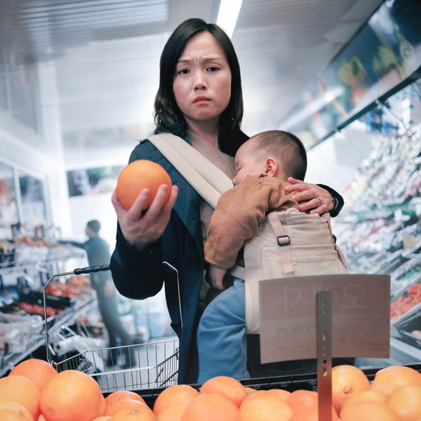 A mother anxiously looking at the price of some fruit while she grocery shops with her sleeping baby.