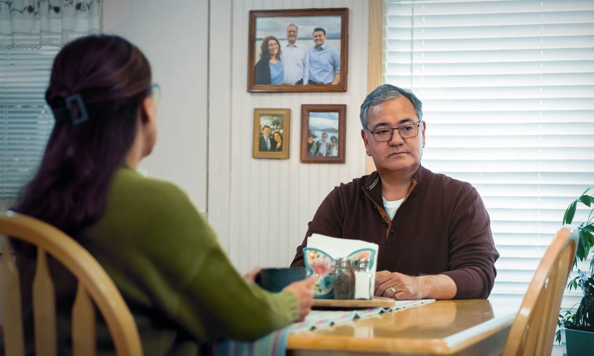 An older couple sitting at a dinner table. The wife stares at family photos of the couple with their son while the husband stares at an empty chair at the table.