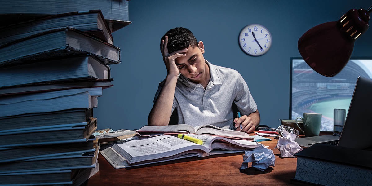 A teenage boy doing his homework late at night while the television is turned on.