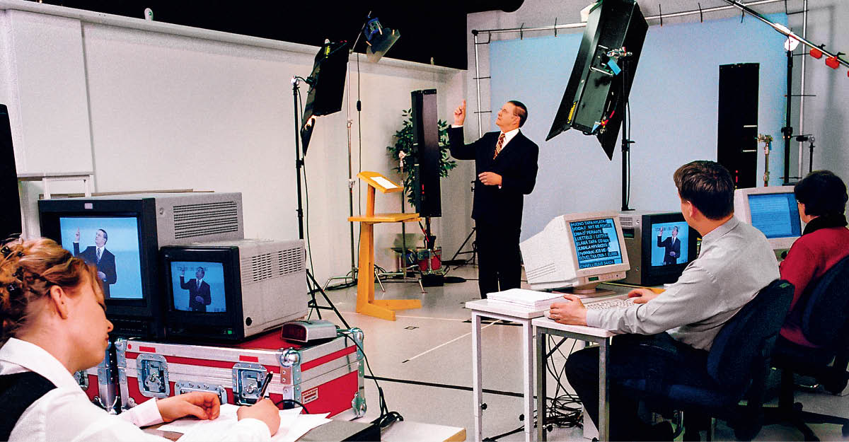 Tapani signing in a studio for a video recording. Three others, including Maire, follow the recording on video monitors.