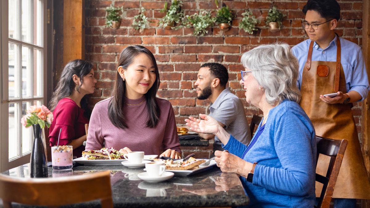 A young sister listening to an older sister’s advice while they eat a meal together at a restaurant.