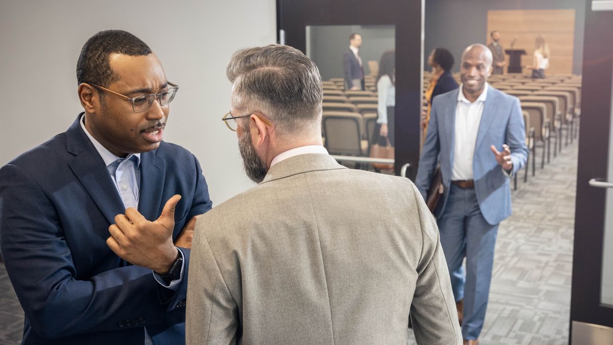 Two brothers conversing with each other at the Kingdom Hall. One of the brothers speaks negatively about another brother who is walking toward them.