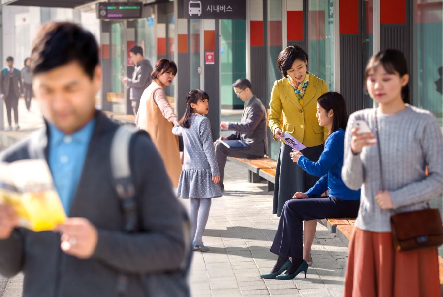 A sister giving a tract to a woman at a bus station while many passersby seem indifferent to her preaching.