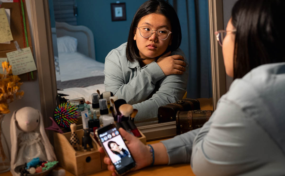 A teenage girl looking at herself in a mirror and comparing her appearance with that of a girl on her smartphone.