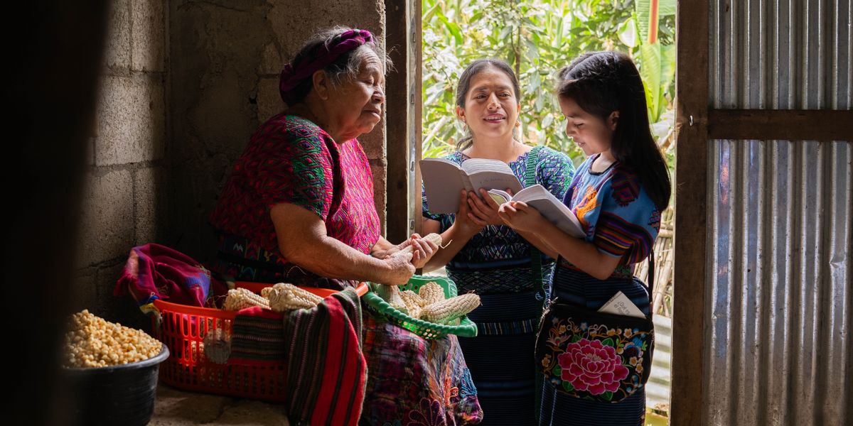 A Witness girl and her mother reading a scripture to a woman who is husking corn in her home.