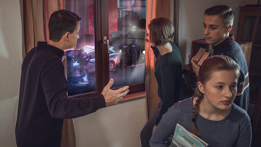 Di saym famili eena di fers picha. Di payrents di luk chroo di windo ahn dehn si di polees deh owtsaida dehn hows, wail di son ahn di daata dehn kaam ahn dehn di goh haid di Baibl bays poblikayshan dehn.