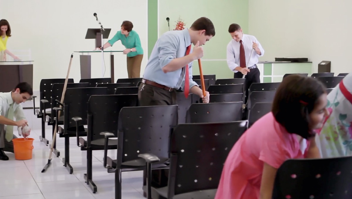 Scéna z videa „Staráme se o místa, kde uctíváme Jehovu“. Muži, ženy i děti uklízí sál Království
