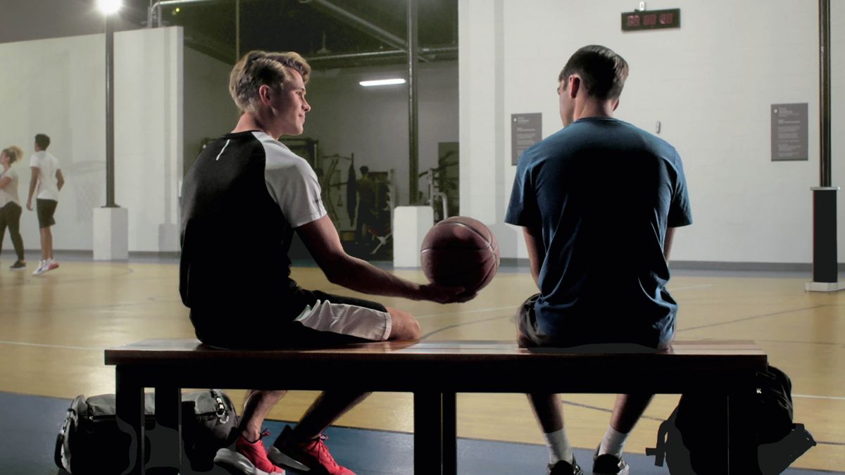 Scéna z videa „‚Všechno má svůj čas‘: Přátelství“. Lucas sedí s Benem na lavičce v tělocvičně a podává mu míč na basket.