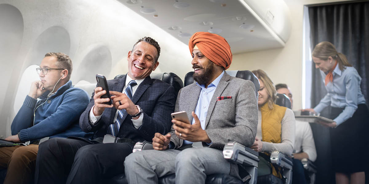 Un hombre blanco y un hombre de la religión sijista sentados juntos en un avión conversando alegremente.