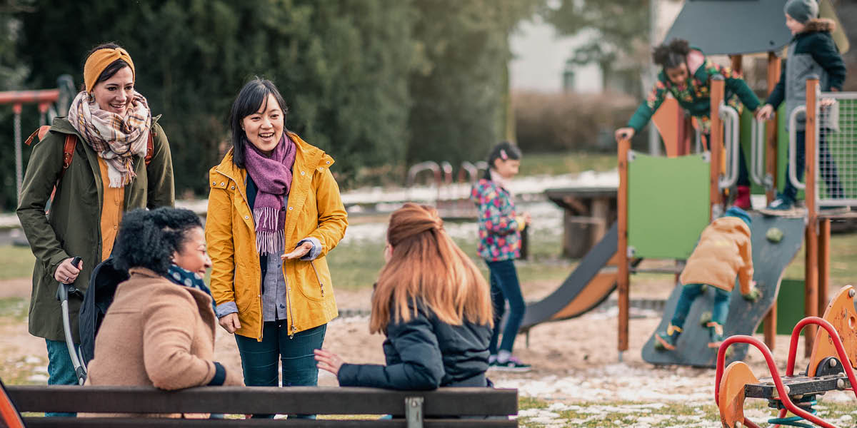Cuatro mujeres de diferentes razas hablando y riéndose mientras sus hijos juegan juntos en el parque.