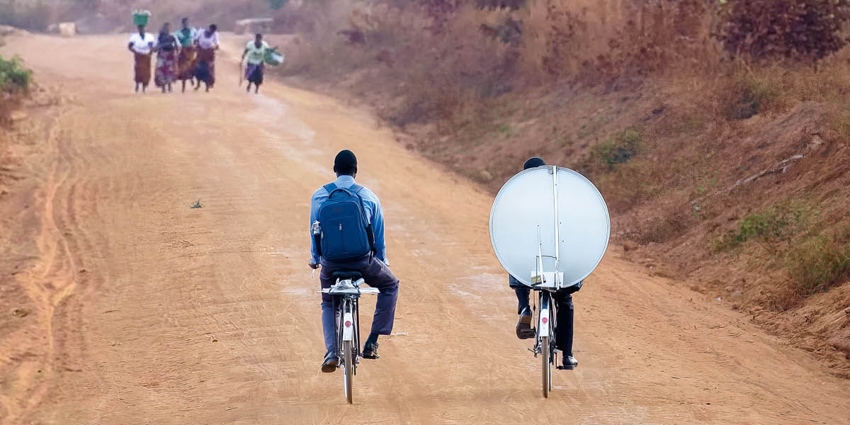 Hermanos en bicicleta en una calle de tierra llevan al Salón del Reino el equipo para ver el canal por satélite.