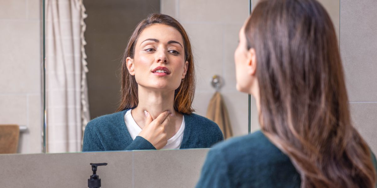 Frente a un espejo, una mujer se toca el cuello para comprobar si tiene algún problema en la tiroides.