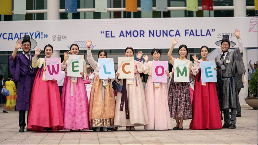 Una imagen del video Las asambleas internacionales “El amor nunca falla”. Un grupo de hermanos coreanos sostiene un cartel de bienvenida y saludan a los que llegan a la asamblea.