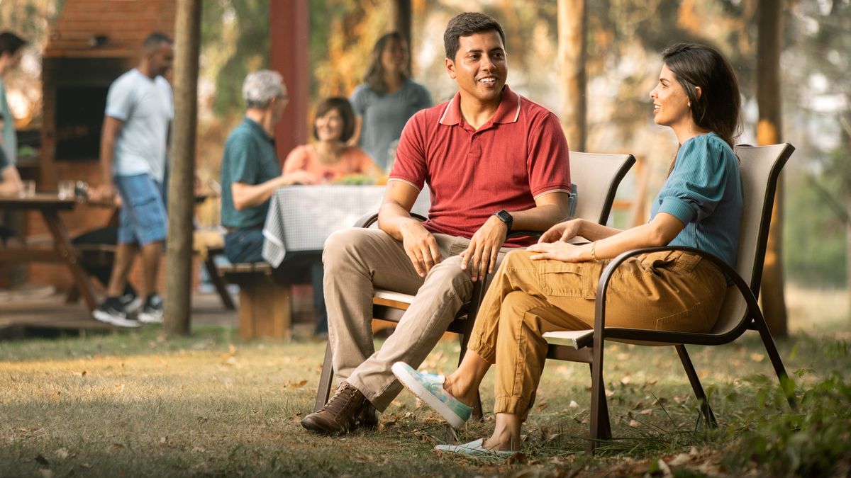 Durante una reunión social, una pareja de novios conversa tranquilamente sentada. Al fondo hay otros hermanos.
