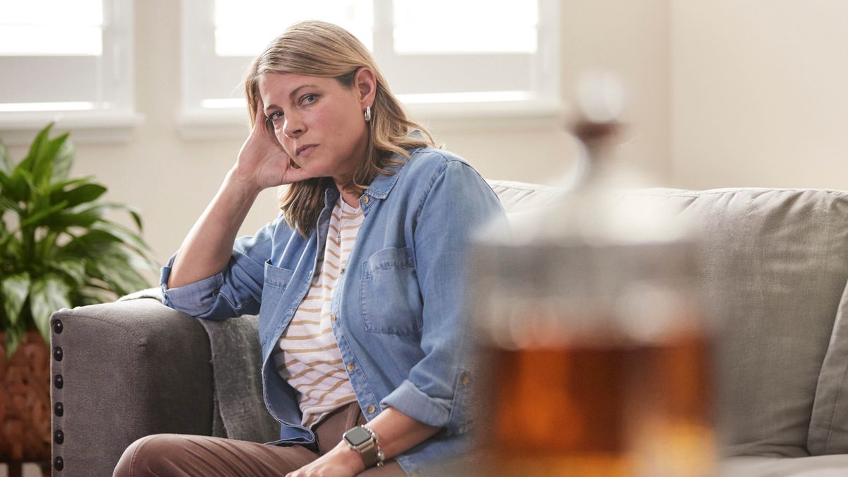 Una hermana sentada en el sofá se queda mirando una botella de alcohol que está al otro lado de la sala.