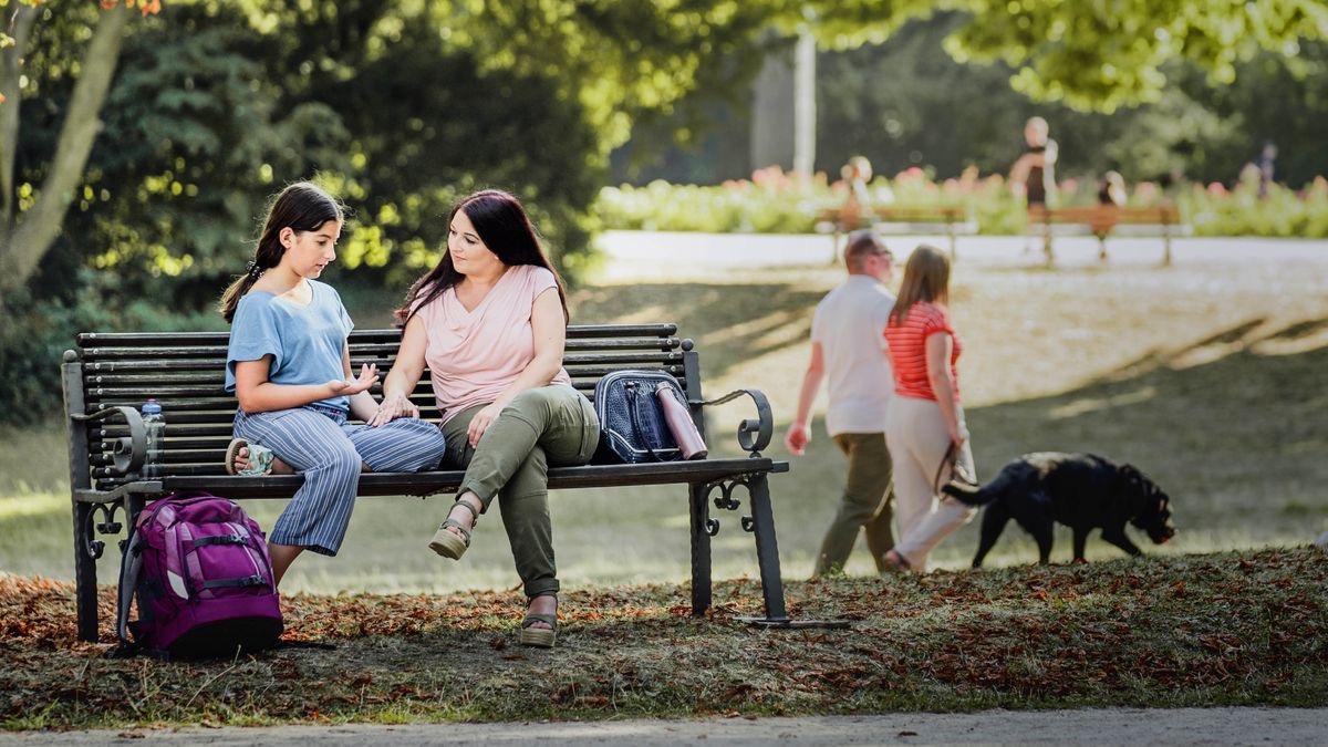 Una jovencita le cuenta a su mamá cómo se siente. Están sentadas en una banca de un parque.