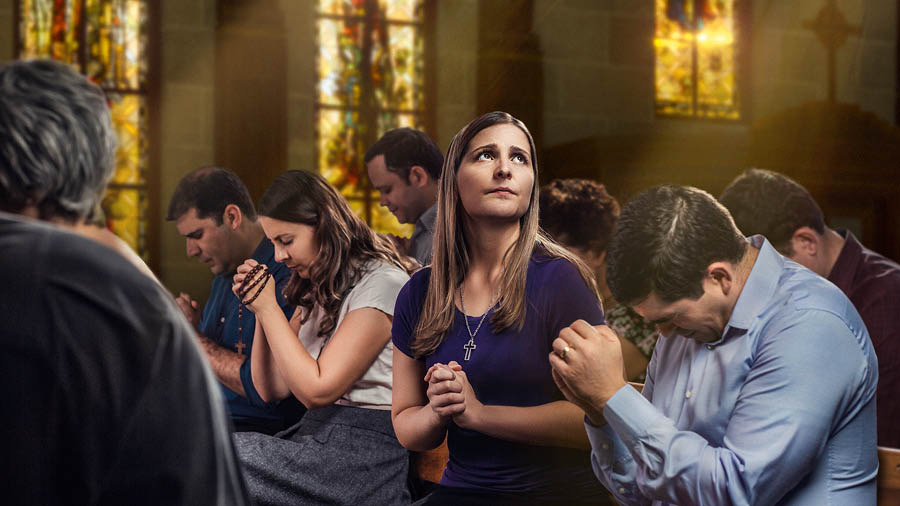 Una mujer en una iglesia ora mirando hacia arriba mientras otras personas inclinan su cabeza para orar.