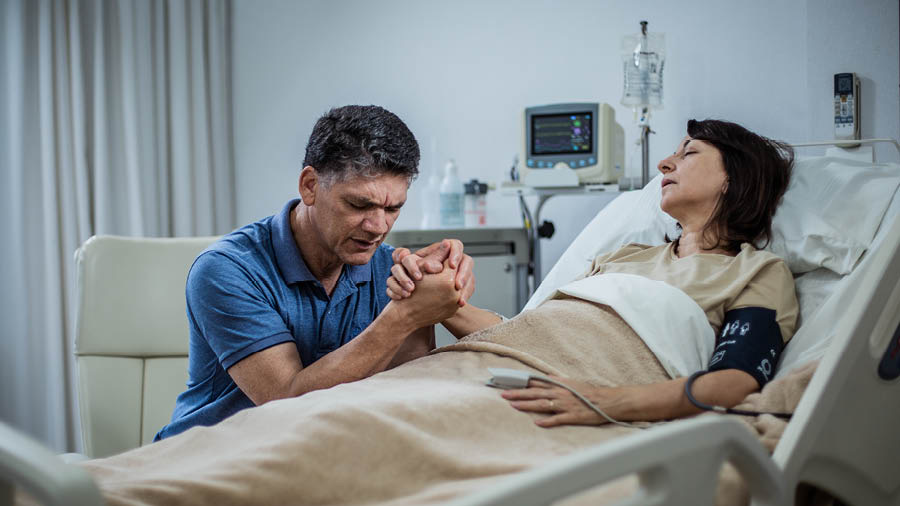 Un esposo ora con su esposa que está en la cama de un hospital.