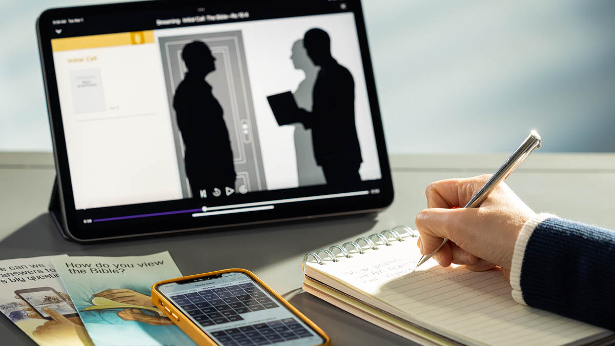En søster sidder og tager notater mens hun ser en video fra midtugemødet med et forslag til en samtale. Ved siden af hendes tablet ligger der nogle traktater og en telefon hvor JW Library er åben.