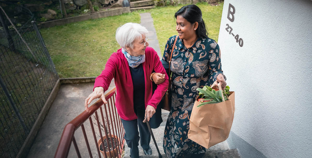 Una mujer de la India ayudando a una anciana de raza blanca a subir la bolsa con las compras por las escaleras.