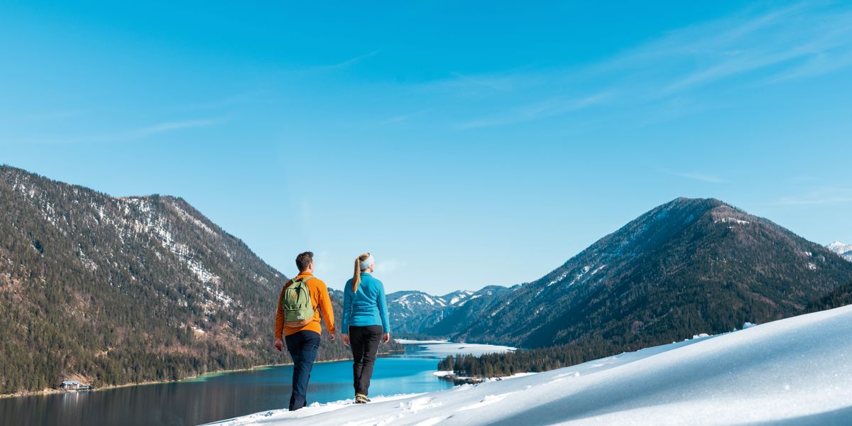 Bajo un cielo azul, un matrimonio camina por una colina nevada mientras mira un lago rodeado de montañas llenas de árboles.