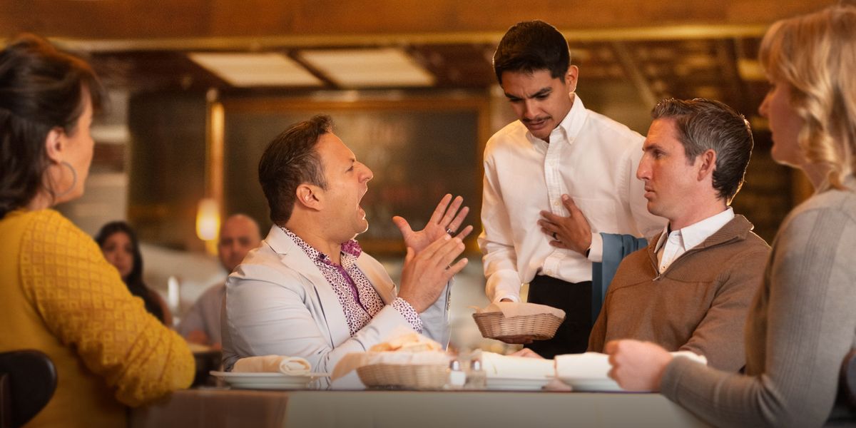 En un restaurante, un hombre le habla muy enojado al camarero o mesero, quien está intentando disculparse. El resto de personas que están en la mesa se ven incómodas por la situación.