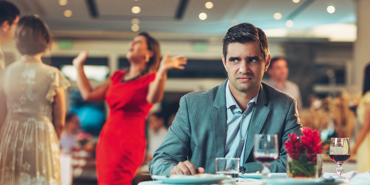 Un hombre, que parece estar molesto, está sentado a la mesa solo mientras su esposa tiene una conversación muy animada con otro matrimonio.