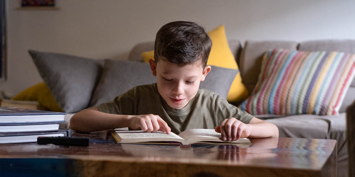 Un niño leyendo un libro. En la mesa hay un control remoto y más libros.