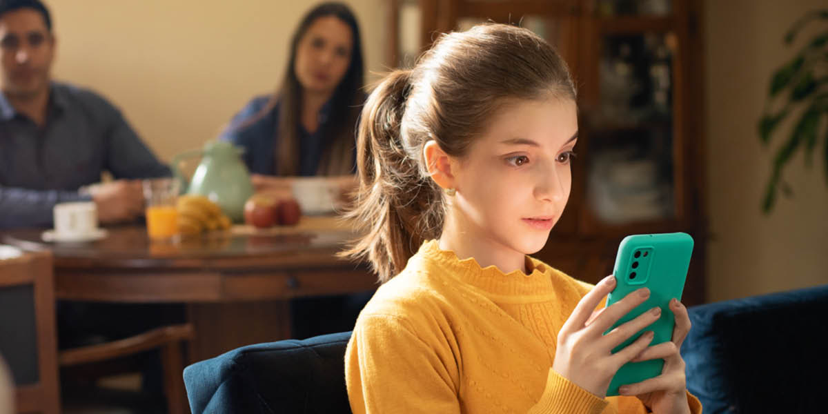 Una chica pegada a su teléfono. Sus padres la miran mientras la esperan sentados a la mesa para desayunar.