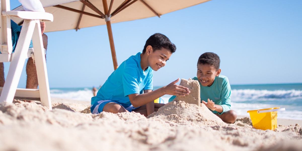 Unos niños construyendo un castillo de arena en la playa mientras su padre está sentado cerca de ellos.
