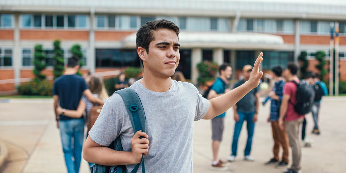 Un joven hace un gesto de desprecio con la mano mientras se va de la escuela. Al fondo hay un grupo de estudiantes.