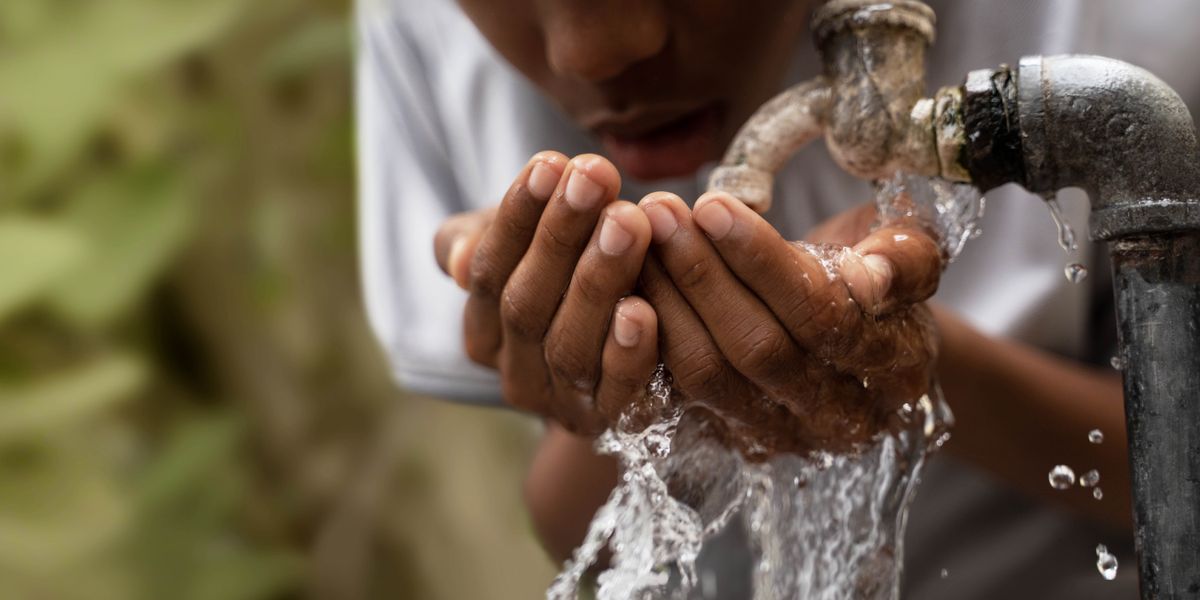 En la calle, un muchacho recoge con sus manos agua de un grifo y se la bebe.