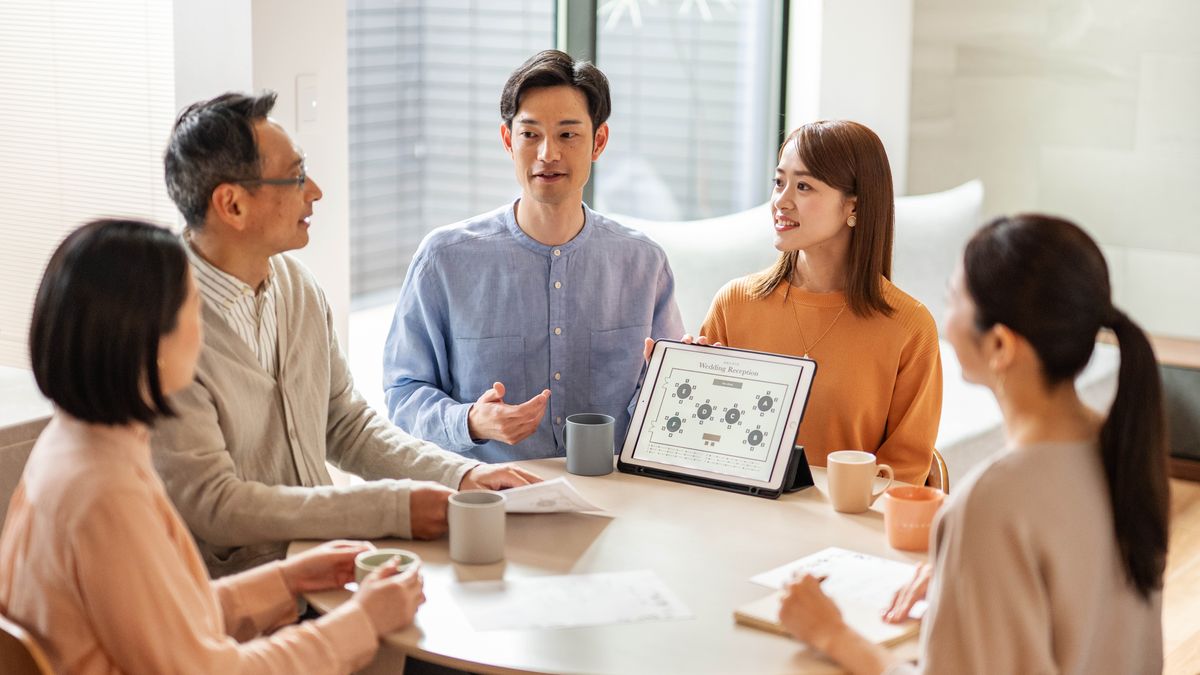 An engaged couple discussing their wedding arrangements with some friends who will be assisting them. The brother holds a tablet and shows the plans for the seating arrangement at the wedding reception.