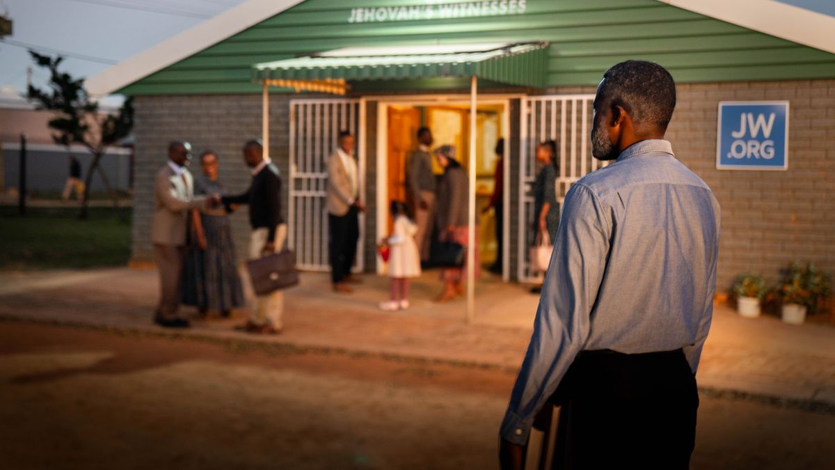 An older man with a Bible in hand, standing a short distance away from a Kingdom Hall. He observes brothers and sisters warmly welcoming one another as they arrive.