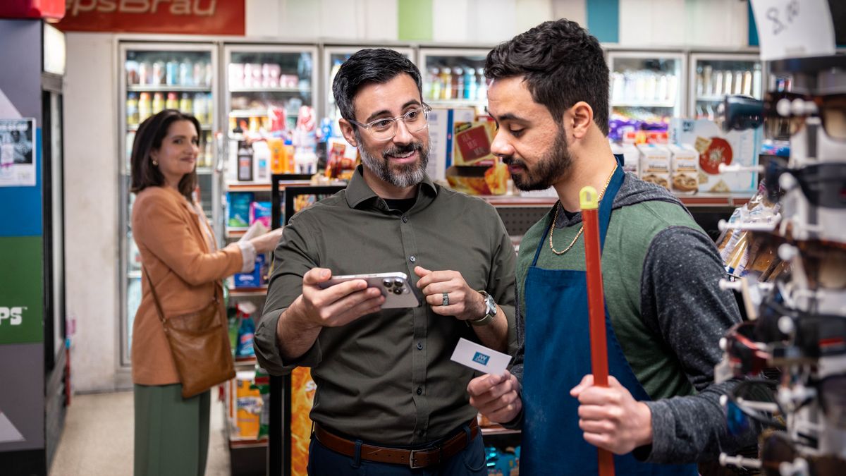 Later, the brother hands a jw.org contact card to an employee at a convenience store and shows him something on his phone. His wife stands nearby, watching with a smile.