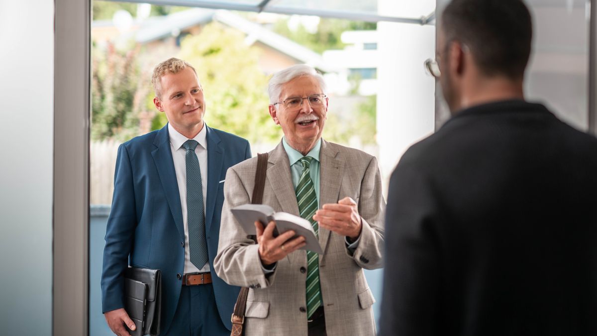The elder and older brother work together in the ministry. The older brother confidently discusses a scripture with a man standing at his door.