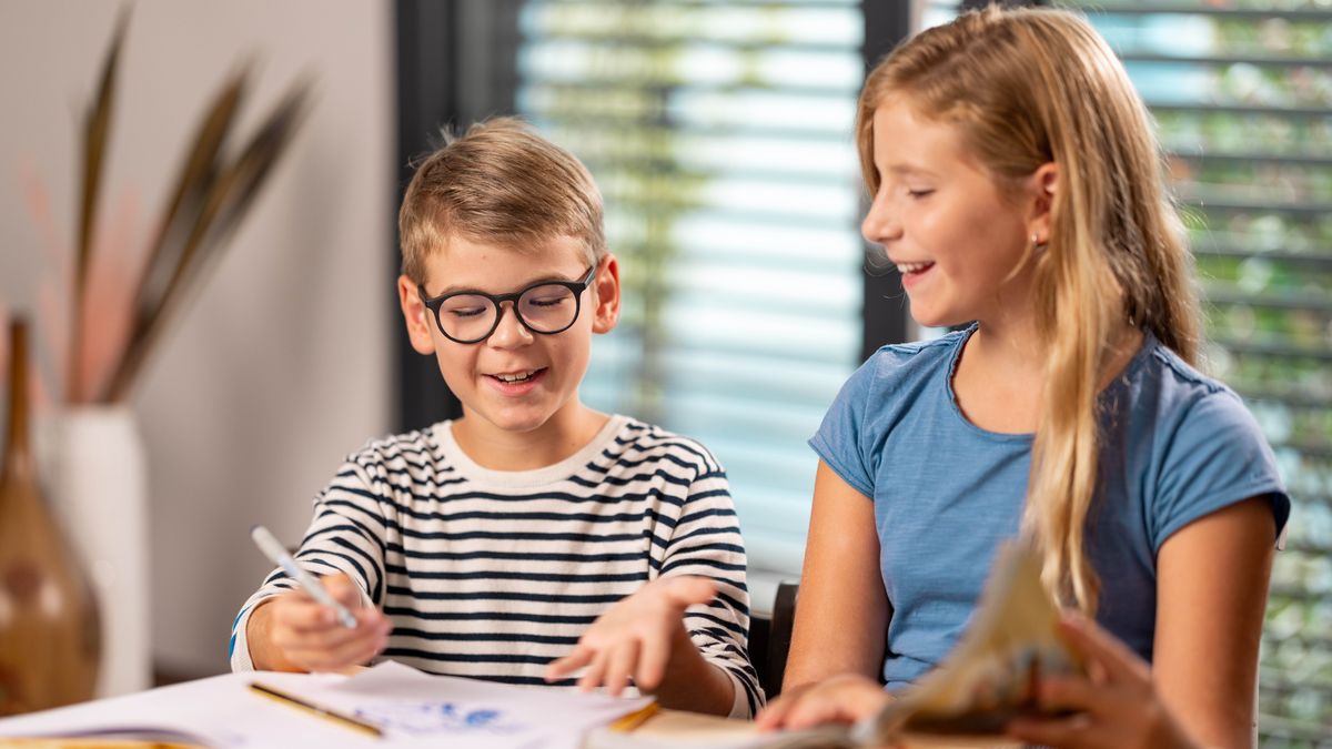 A young sister sits with her younger brother and reads from a book while he draws pictures of Bible scenes.