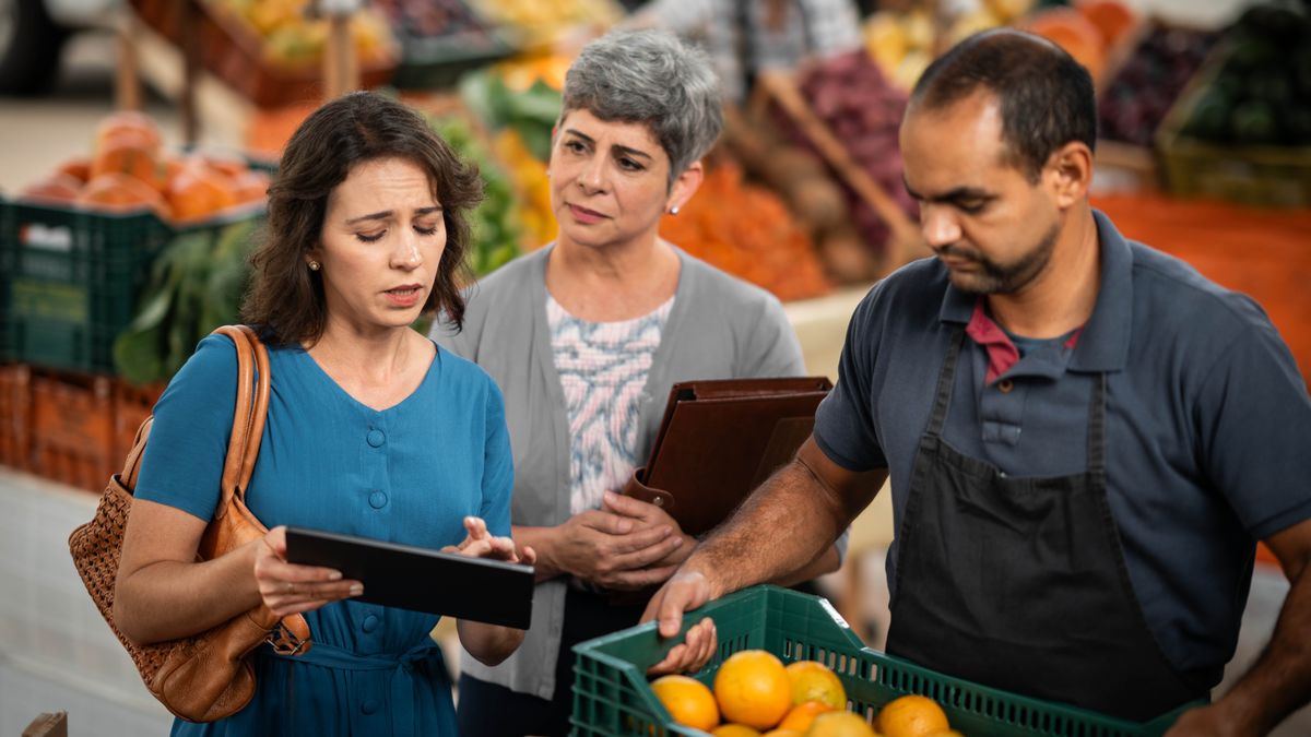 A mature sister observing another sister struggling to give a witness to a man working at a market.