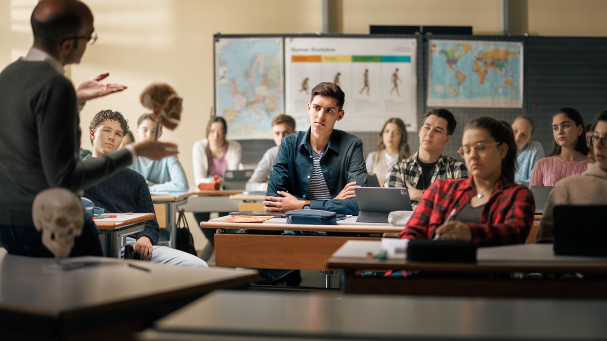 En un aula, un adolescente escucha con respeto mientras su profesor de ciencias sostiene un cráneo y se dirige a él. En la parte de atrás, hay un cuadro que muestra las fases de la evolución humana.