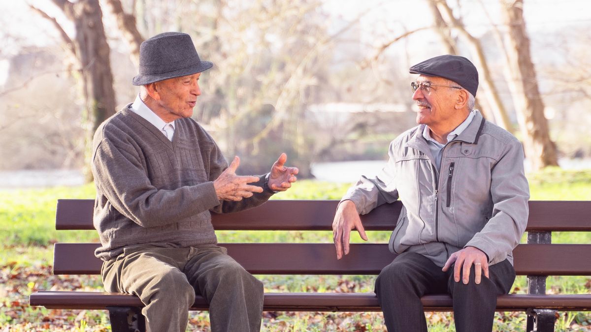 Dans un jardin public, un frère âgé et un autre homme discutent, assis sur un banc.