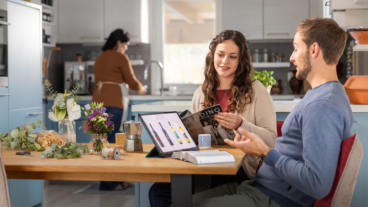 Des fiancés sont en train de décider s’ils serviront ou non de l’alcool à leur réception de mariage. Sur la table se trouvent une bible ouverte ainsi qu’une tablette affichant un choix de bouteilles de vin.
