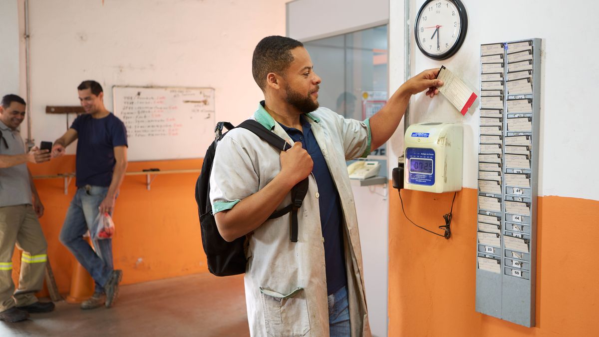 Veli saapuu työpaikalleen aamulla kello 7:29. Taustalla on muita työntekijöitä, jotka katsovat jotain puhelimelta.