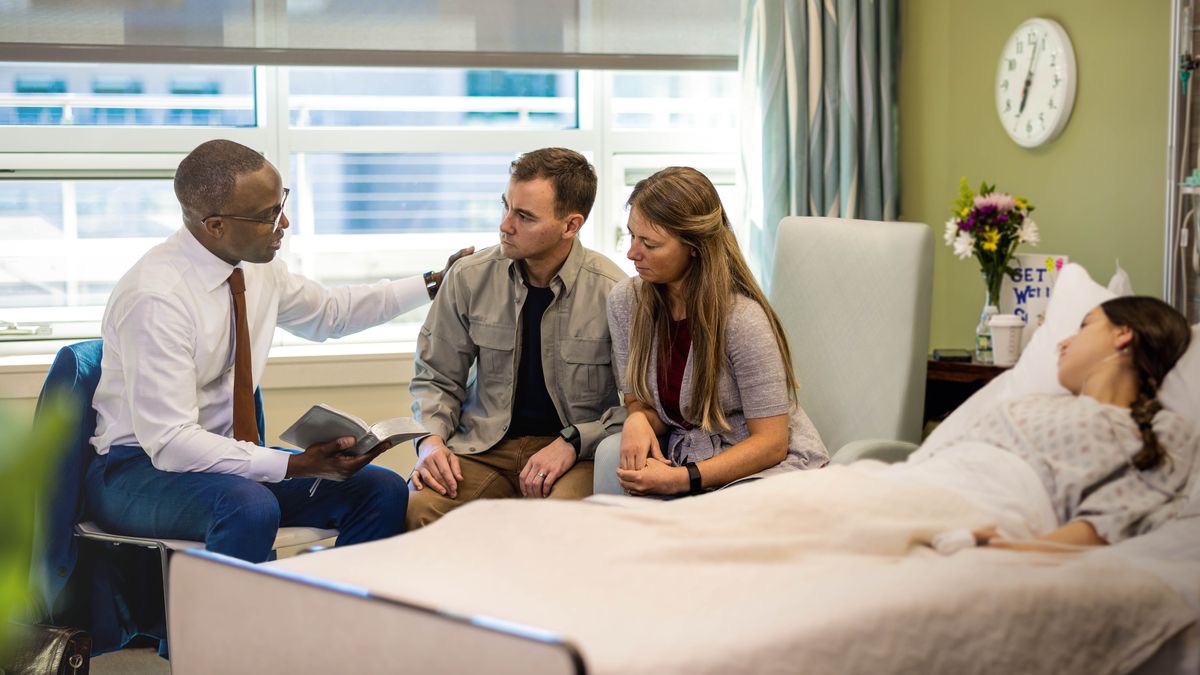 An elder using the Bible to comfort a couple. A young woman lies in a hospital bed next to them.