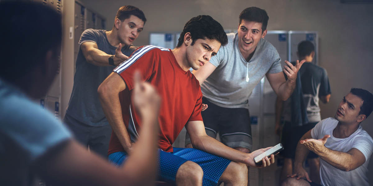 Un adolescente, nun vestiario, sostendo un teléfono móbil na man mentres outros rapaces exercen presión para que tome unha decisión.