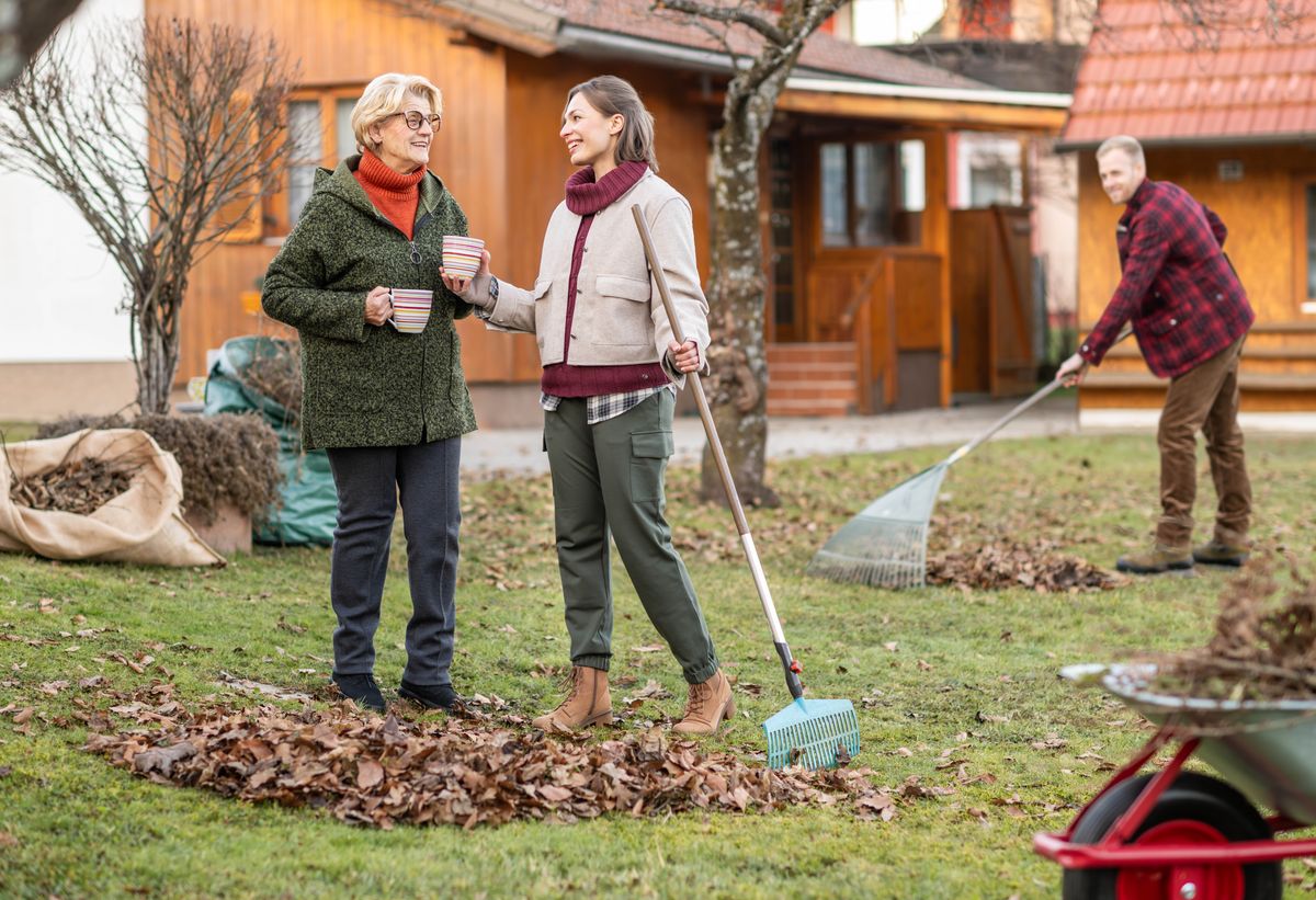 Ein Ehepaar hilft einer älteren Frau, das Laub aus ihrem Garten zu entfernen; sie bringt ihnen ein heißes Getränk.