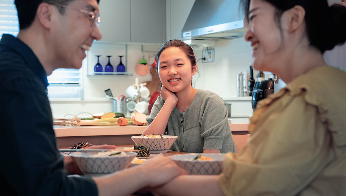 Eine Tochter im Teenageralter beobachtet lächelnd ihre Eltern beim Essen. Der Vater hält die Hand der Mutter und sie lachen zusammen.