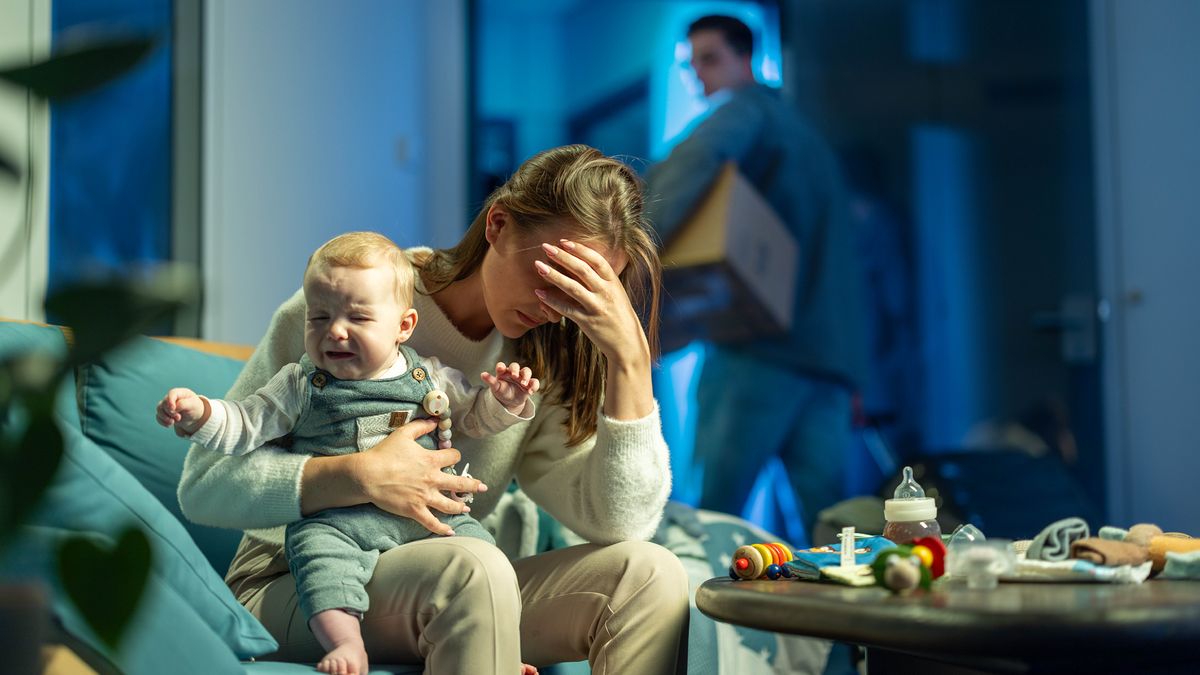 Die Frau aus dem vorherigen Bild hat ein schreiendes Baby auf dem Schoß und vergräbt ihren Kopf in ihrer Hand. Der Mann trägt eine Kiste mit persönlichen Dingen und zieht aus.