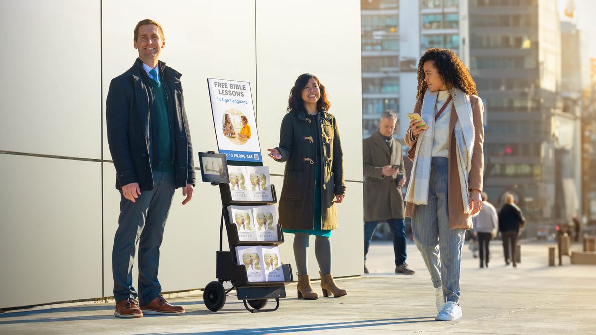 Ein Ehepaar steht an einem Trolley mit dem Poster „Kostenloser Bibelkurs“. Eine Passantin schaut es sich an.