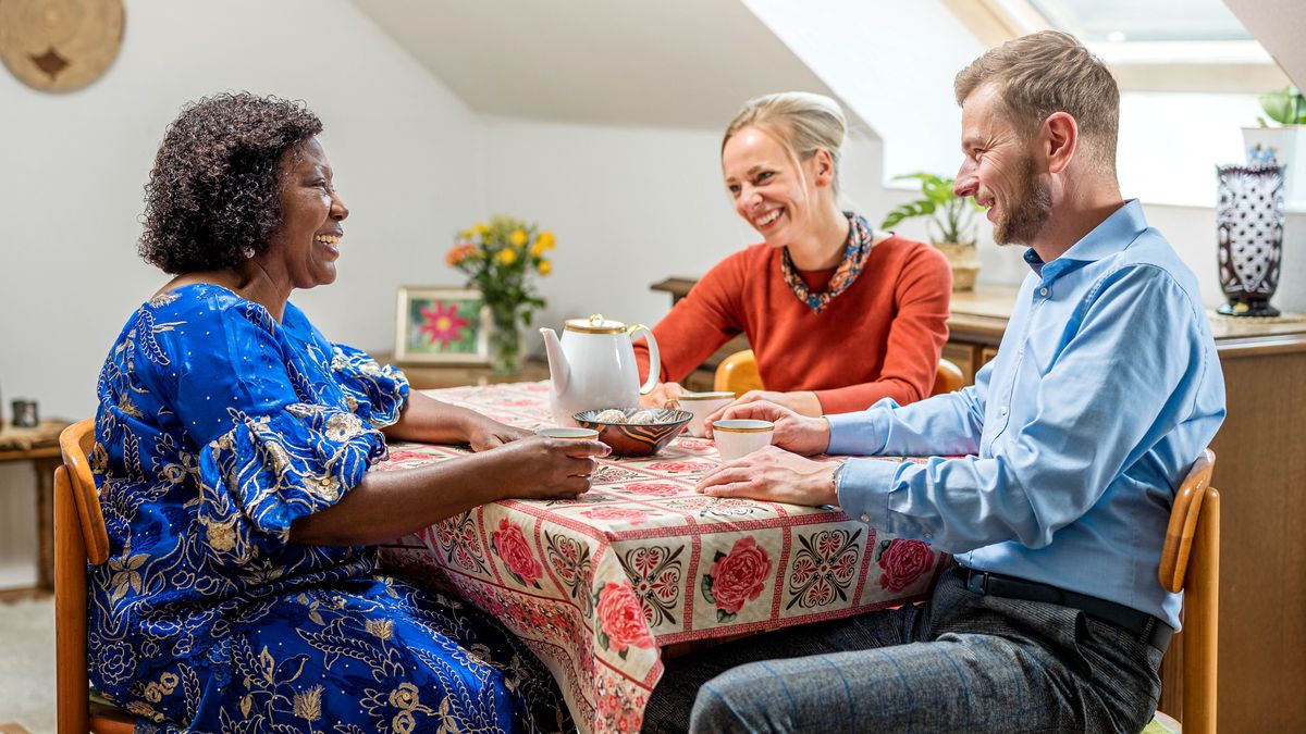 Ein junges Ehepaar besucht eine ältere Schwester und sitzt mit ihr am Kaffeetisch.