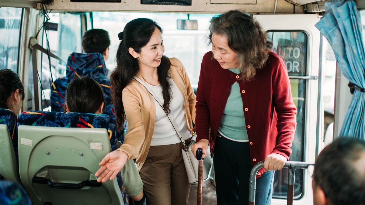 Eine Schwester bietet einer älteren Frau im Bus ihren Sitzplatz an.